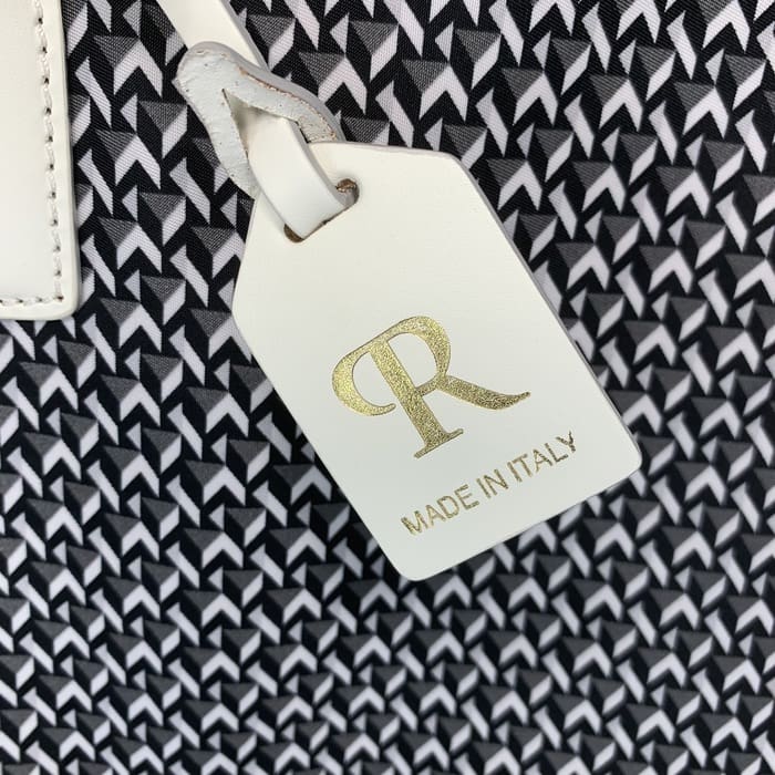 Белая кожаная бирка ROBERTA PIERI с буквами «R» золотого цвета и надписью «MADE IN ITALY» свисает на фоне черно-белой геометрической ткани.