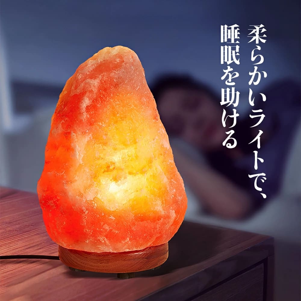 天然岩塩ランプ【サイズ：SS】— ほっと落ち着く、やさしい間接照明がベッドサイドテーブルに置かれ、部屋を柔らかく照らし出し、そばで誰かが安らかに眠る中、穏やかな雰囲気を醸し出しています。右側には、このランプの優しい光が安らかな眠りをサポートすることを説明する日本語のテキストが掲載されています。.